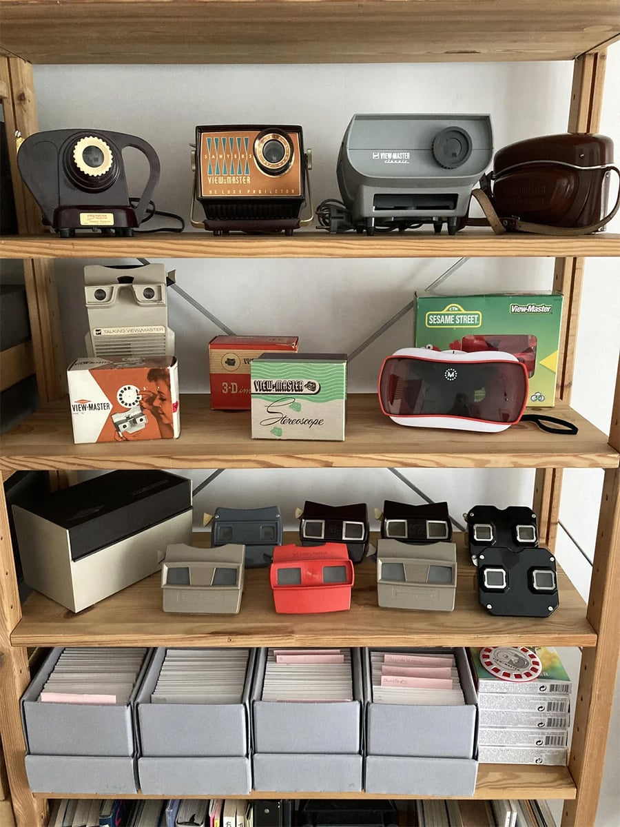 A wooden shelf displays various vintage and modern View-Master viewers, reels, and boxes, along with other retro stereoscopic viewers arranged neatly on multiple shelves.