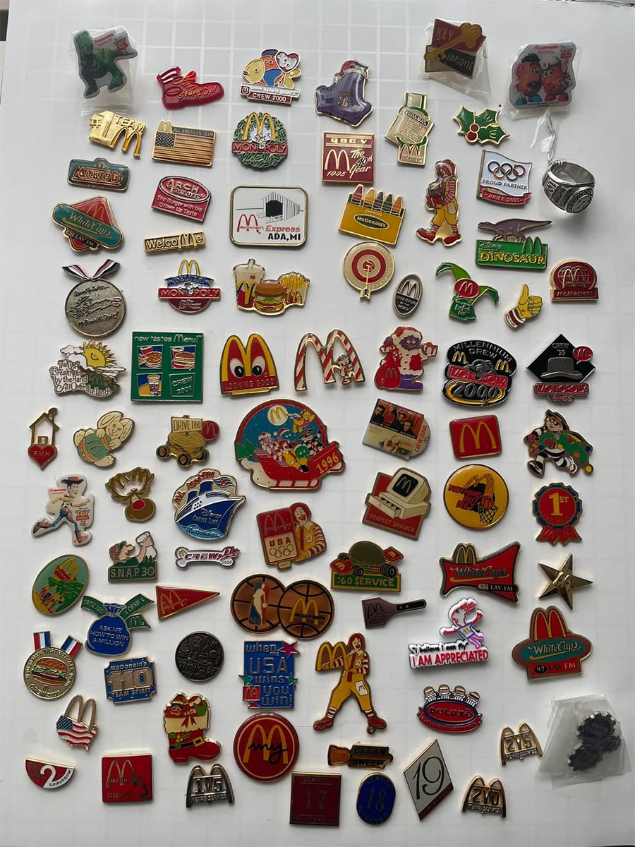 A collection of colorful McDonald’s pins arranged on a white grid, featuring logos, mascots, Olympic themes, and various promotional designs.