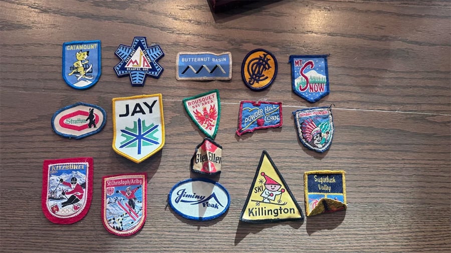 Fifteen colorful ski resort patches are arranged on a wooden surface, with various shapes, text, and ski-themed logos representing different locations.