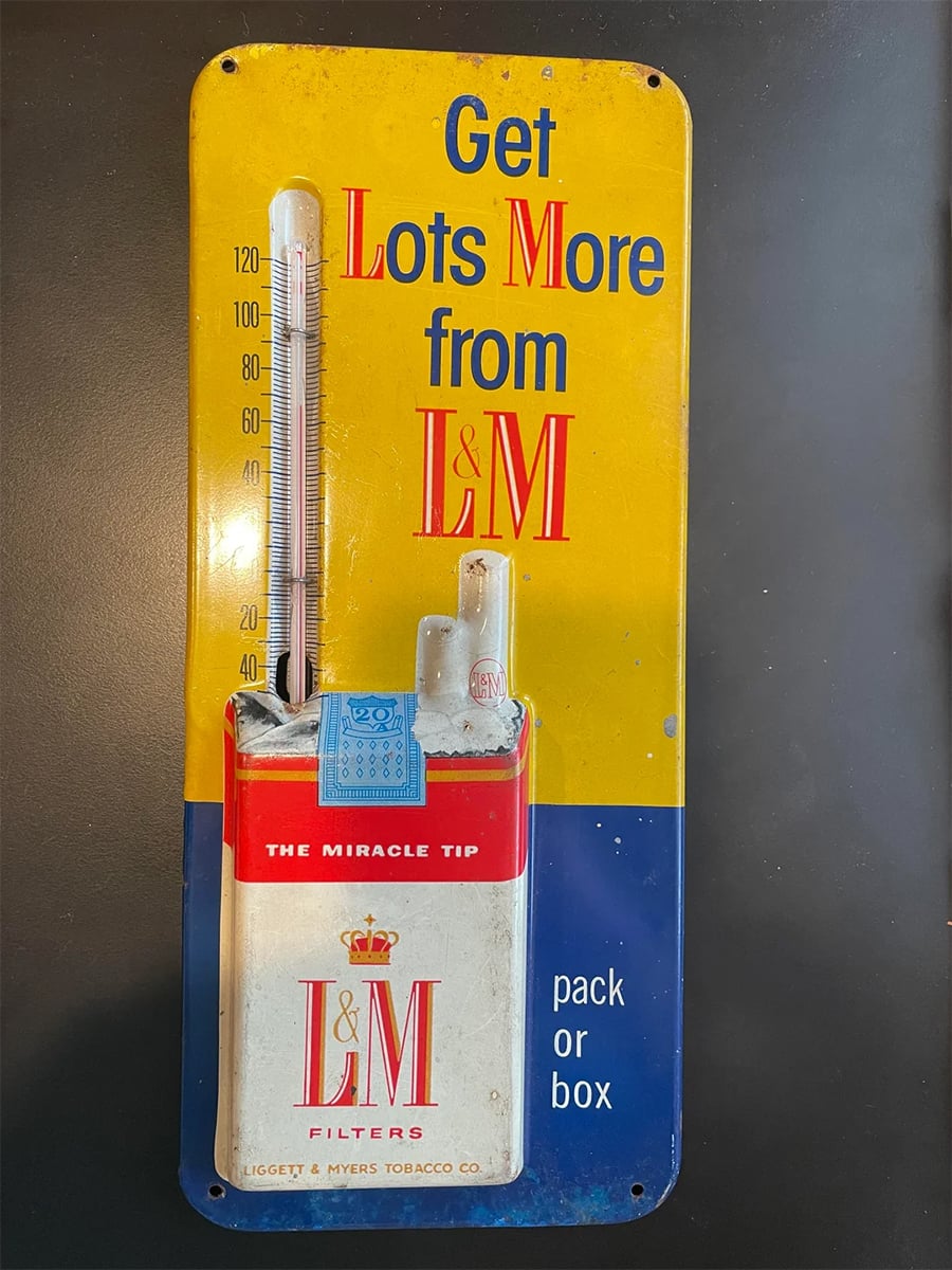 A vintage L&M cigarette thermometer sign reads “Get Lots More from L&M,” featuring a red and white cigarette pack and a vertical thermometer on a blue and yellow background.