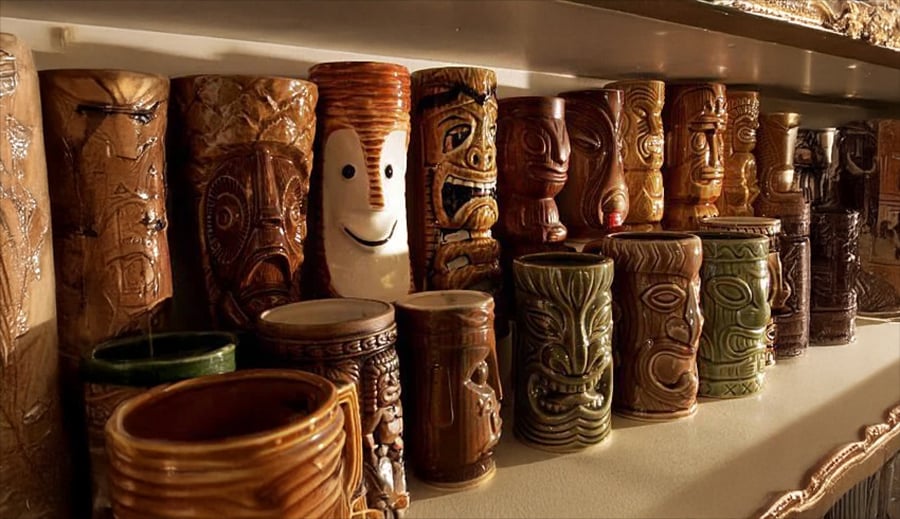 A collection of colorful ceramic tiki mugs with various faces and patterns is displayed on a shelf under warm lighting.