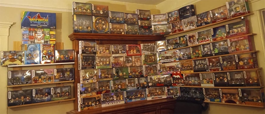Shelves filled with collectible figurines in display boxes, neatly arranged in rows and columns along two corner walls; additional boxed collectibles and posters are visible on the left side.