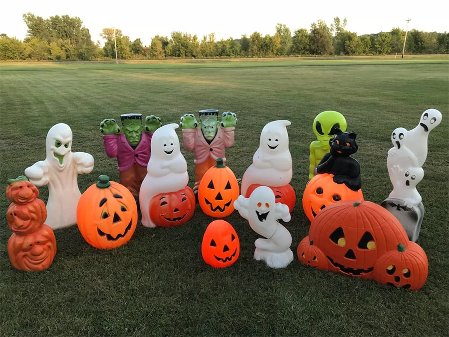 Plastic Halloween decorations, including pumpkins, ghosts, black cat, Frankenstein monsters, and a green alien, are arranged on a grassy lawn with trees in the background.