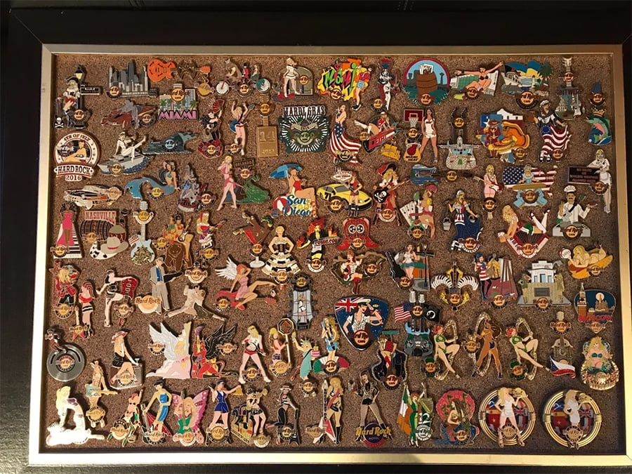 A framed corkboard displays a large, colorful collection of Hard Rock Cafe guitar pins and memorabilia pins, many featuring women, flags, and landmarks from various cities around the world.