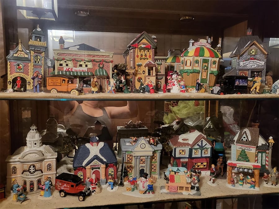 Miniature ceramic village display in a glass cabinet, featuring tiny houses, shops, people, cars, and festive decorations on two shelves. A reflection of a person is visible in the glass behind the scene.