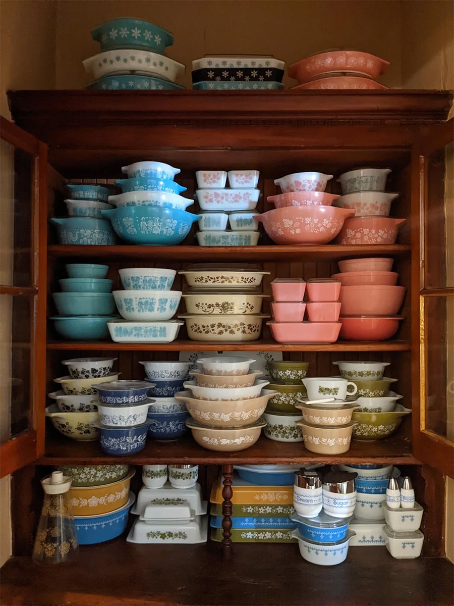 A wooden cabinet holds neatly stacked vintage Pyrex dishes in various shapes, sizes, and pastel colors, including blue, pink, green, and white, with floral and geometric patterns.