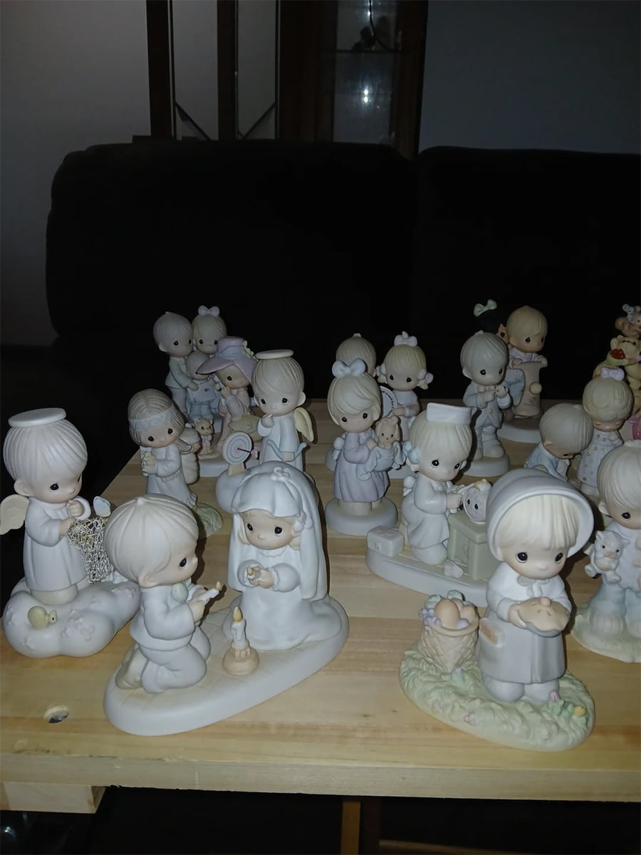 A collection of Precious Moments porcelain figurines is arranged on a wooden table, featuring various angelic and childlike characters in pastel colors.
