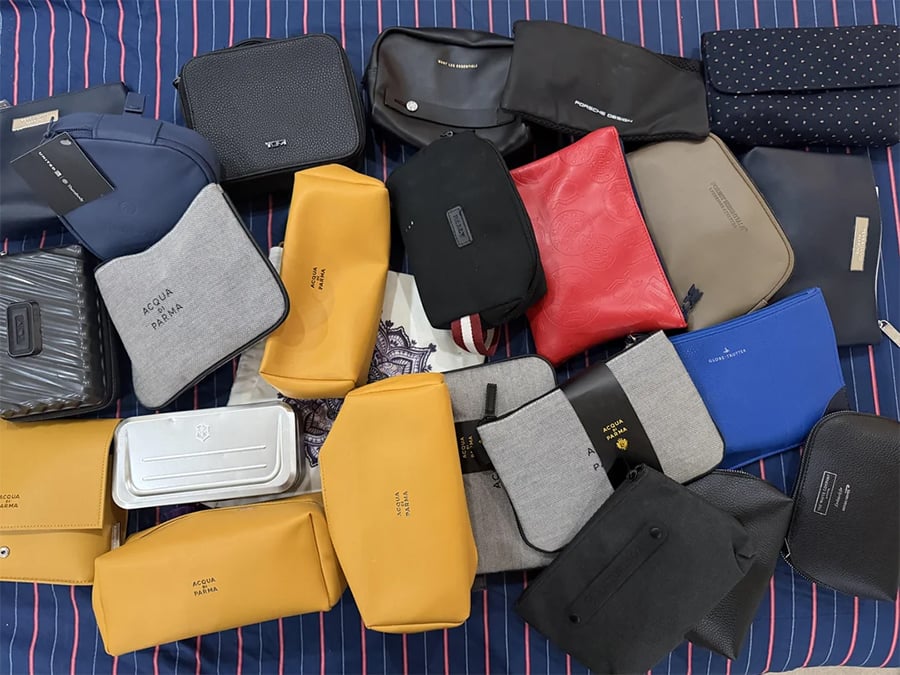 A collection of assorted travel toiletry bags in various colors and shapes is arranged on a blue striped surface. Some bags are yellow, black, gray, blue, or red with different textures and branding.