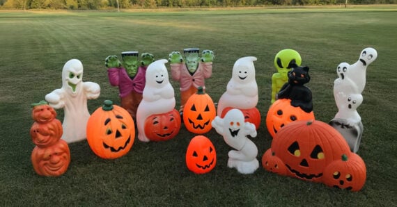 A group of Halloween decorations, including pumpkins, ghosts, a black cat, and green Frankenstein heads, are arranged on grass with trees in the background.