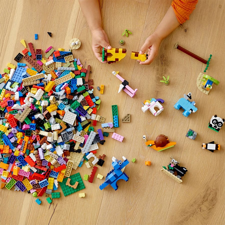 A child’s hands build with colorful LEGO bricks on a wooden floor, with a large pile of assorted pieces and several small completed animal figures nearby.