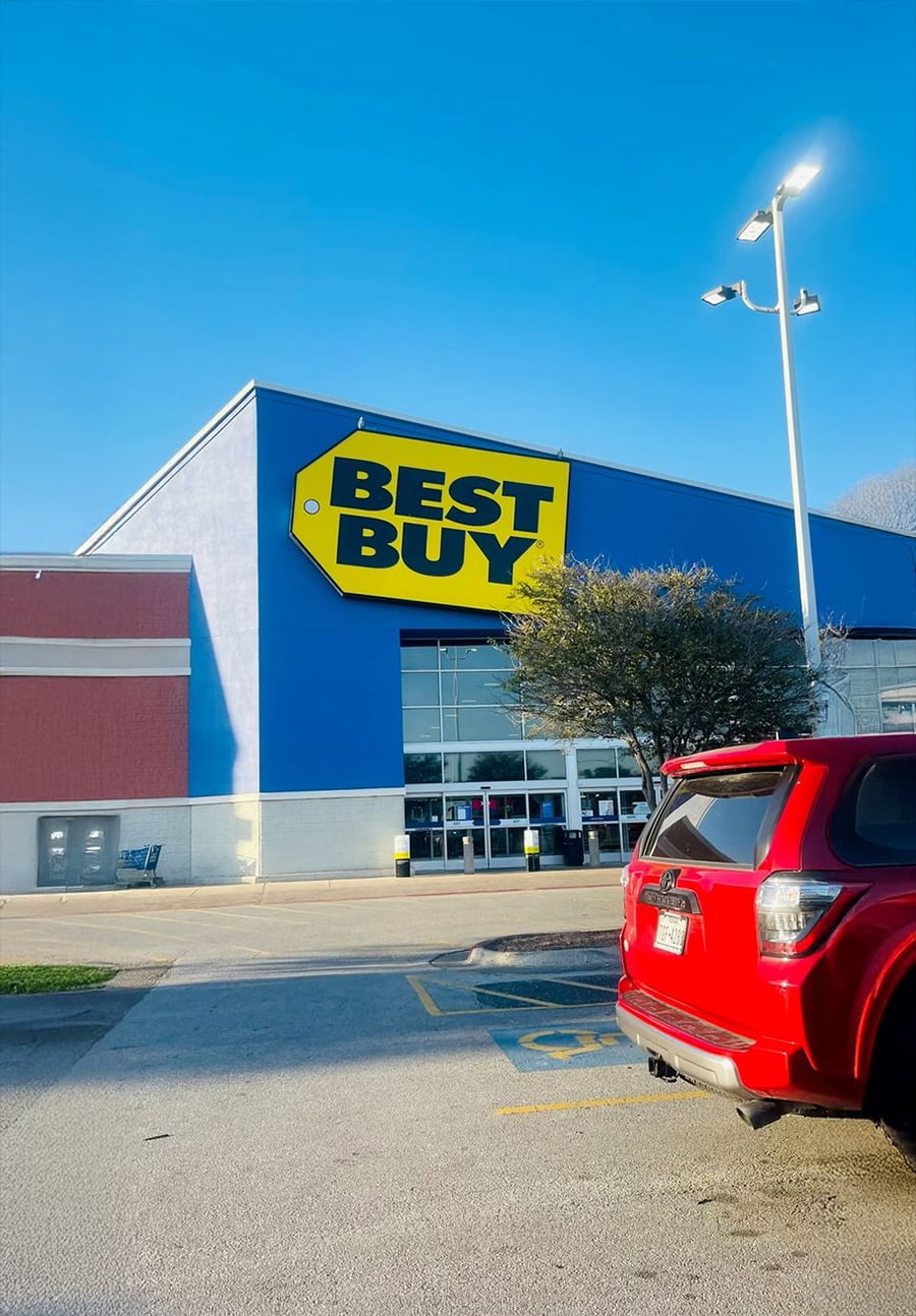 A Best Buy store with a large yellow sign, a tree, and a red SUV parked in front on a sunny day.