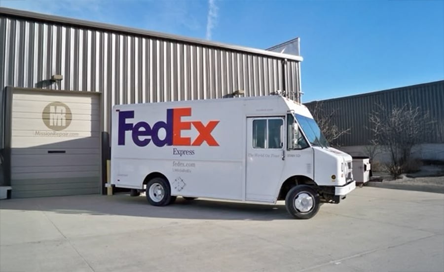 A white FedEx Express delivery truck is parked outside a warehouse loading dock on a sunny day, with a metal roll-up door and industrial building in the background.