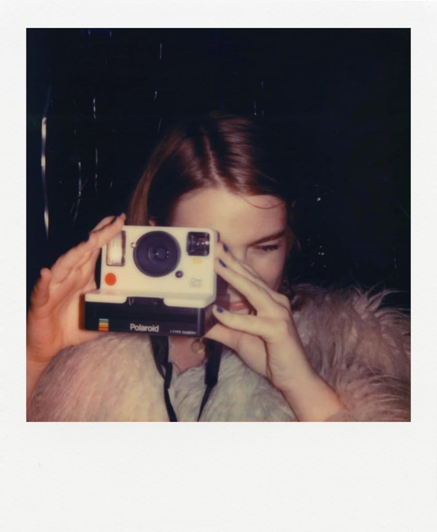 A person with light brown hair holds a vintage Polaroid camera up to their face, preparing to take a photo. The background is dark and they are wearing a light-colored, furry jacket.