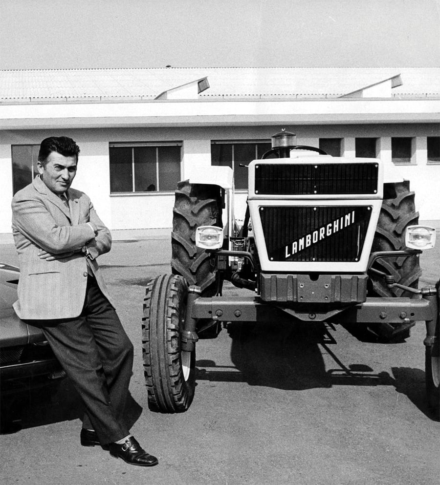 A man in a suit leans against a Lamborghini tractor parked outside a building with large windows on a sunny day.