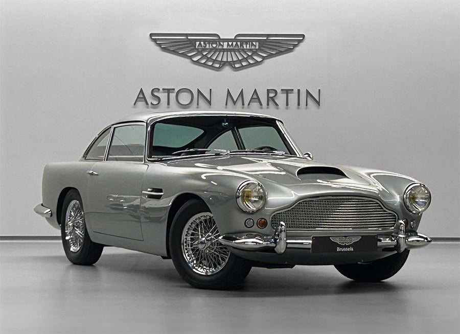 A classic silver Aston Martin coupe is displayed in a showroom with the Aston Martin logo and name on the wall behind it. The car features wire-spoke wheels, round headlights, and a vintage design.