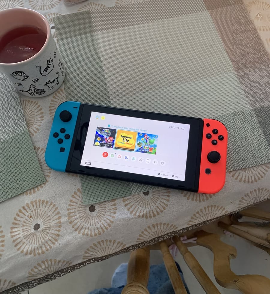 A Nintendo Switch with red and blue Joy-Cons rests on a dining table, showing game icons. A white mug with cat illustrations and a drink inside sits nearby.