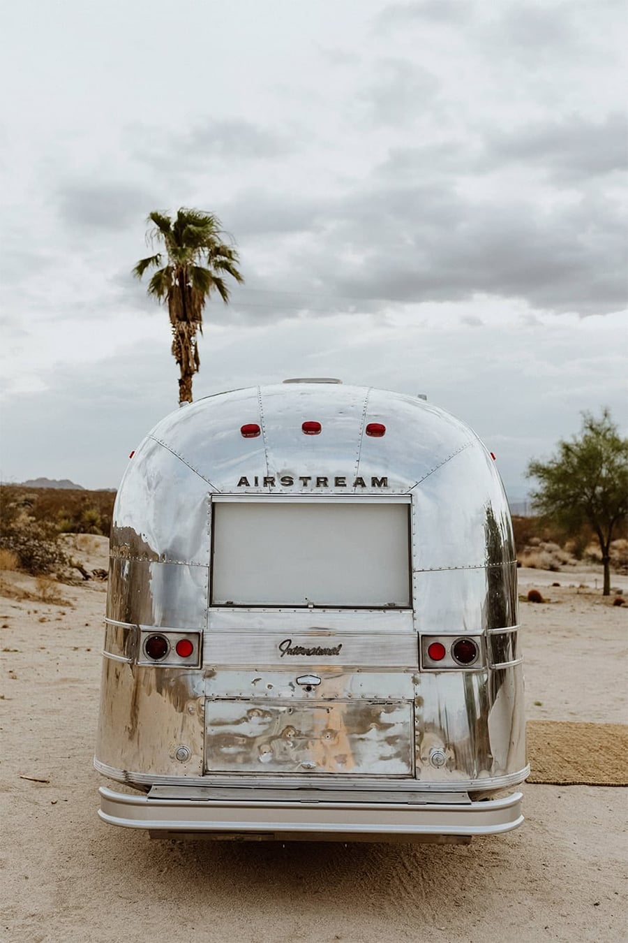 A shiny silver Airstream trailer parked in a sandy desert landscape with a palm tree, sparse vegetation, and a cloudy sky in the background.