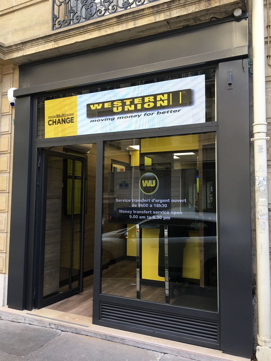 Western Union storefront with glass doors, yellow and black signage, and hours posted in French and English. The sign says, “moving money for better.” The building has a stone exterior with a small balcony above.