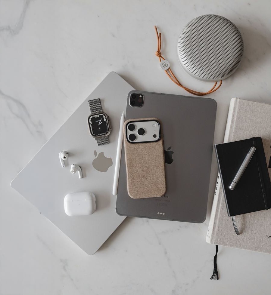 Apple devices including a MacBook, iPad, iPhone, Apple Watch, AirPods, a silver pen, two notebooks, and a round portable speaker arranged on a white marble surface.