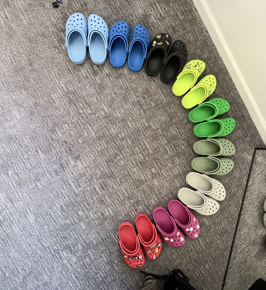 A semicircle of colorful Crocs is arranged on a gray carpet. The shoes are organized by color, ranging from blue and green to black, red, purple, white, and beige.