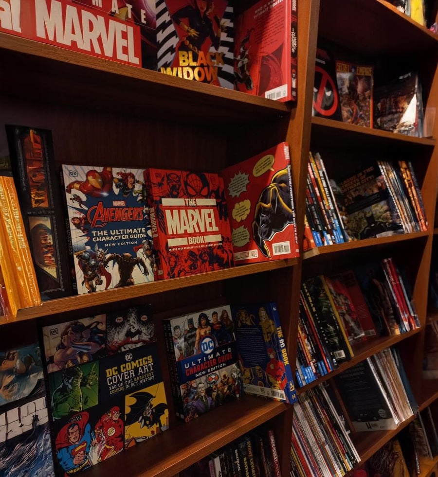 A wooden bookshelf filled with colorful Marvel and DC comic books and graphic novels, including titles like “The Marvel Book” and “The Avengers Ultimate Character Guide.”
