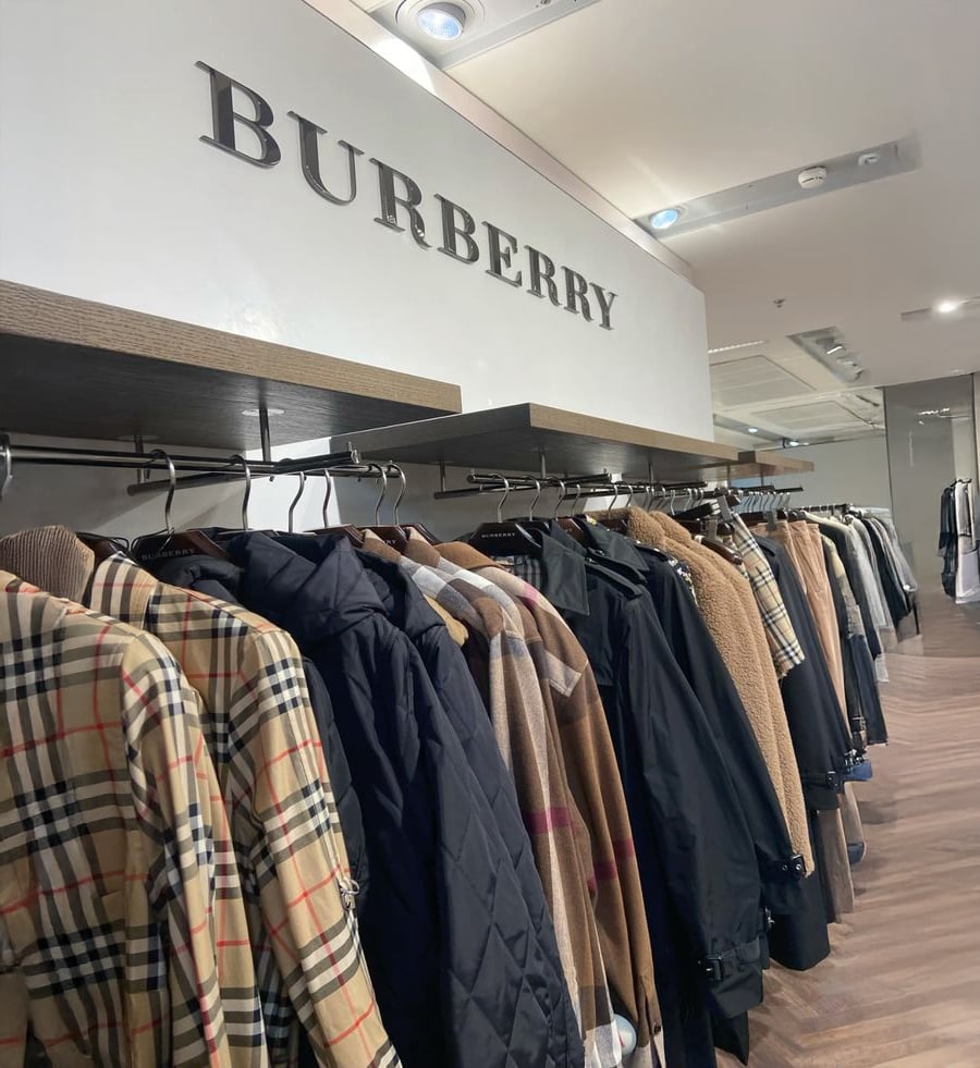 Clothing rack filled with Burberry coats and jackets, including classic checkered patterns, displayed under a large Burberry sign in a modern retail store.