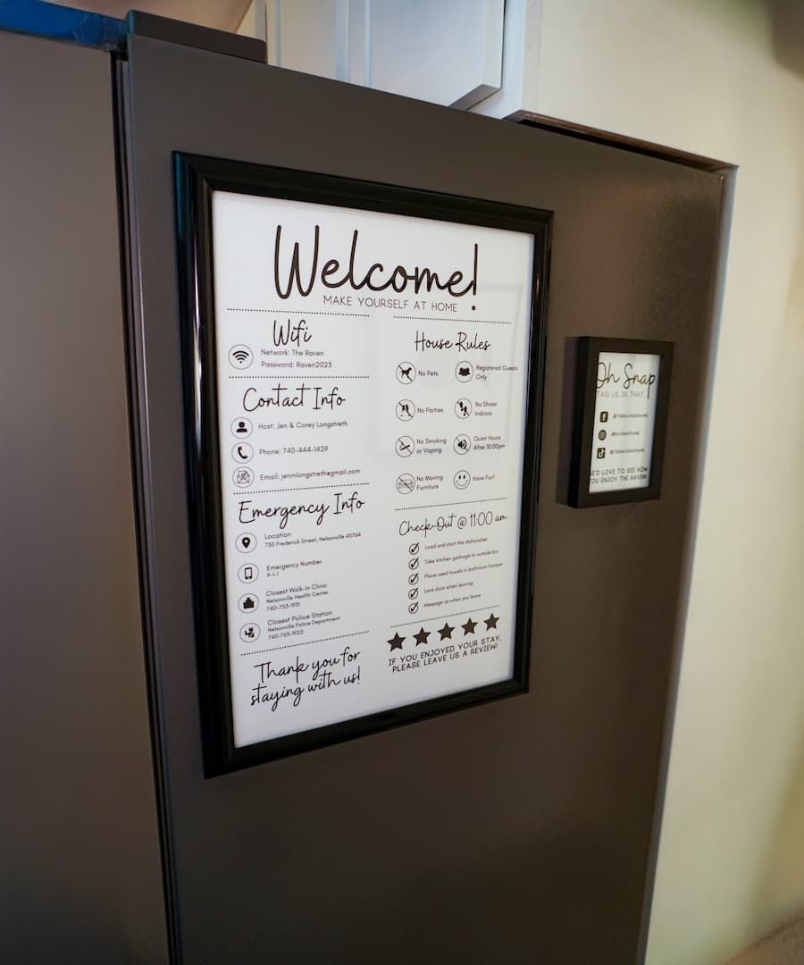 Framed welcome sign with house rules, WiFi info, contact, and emergency info hangs on a dark wall. A smaller framed sign is nearby. Text and icons are visible but not fully readable.