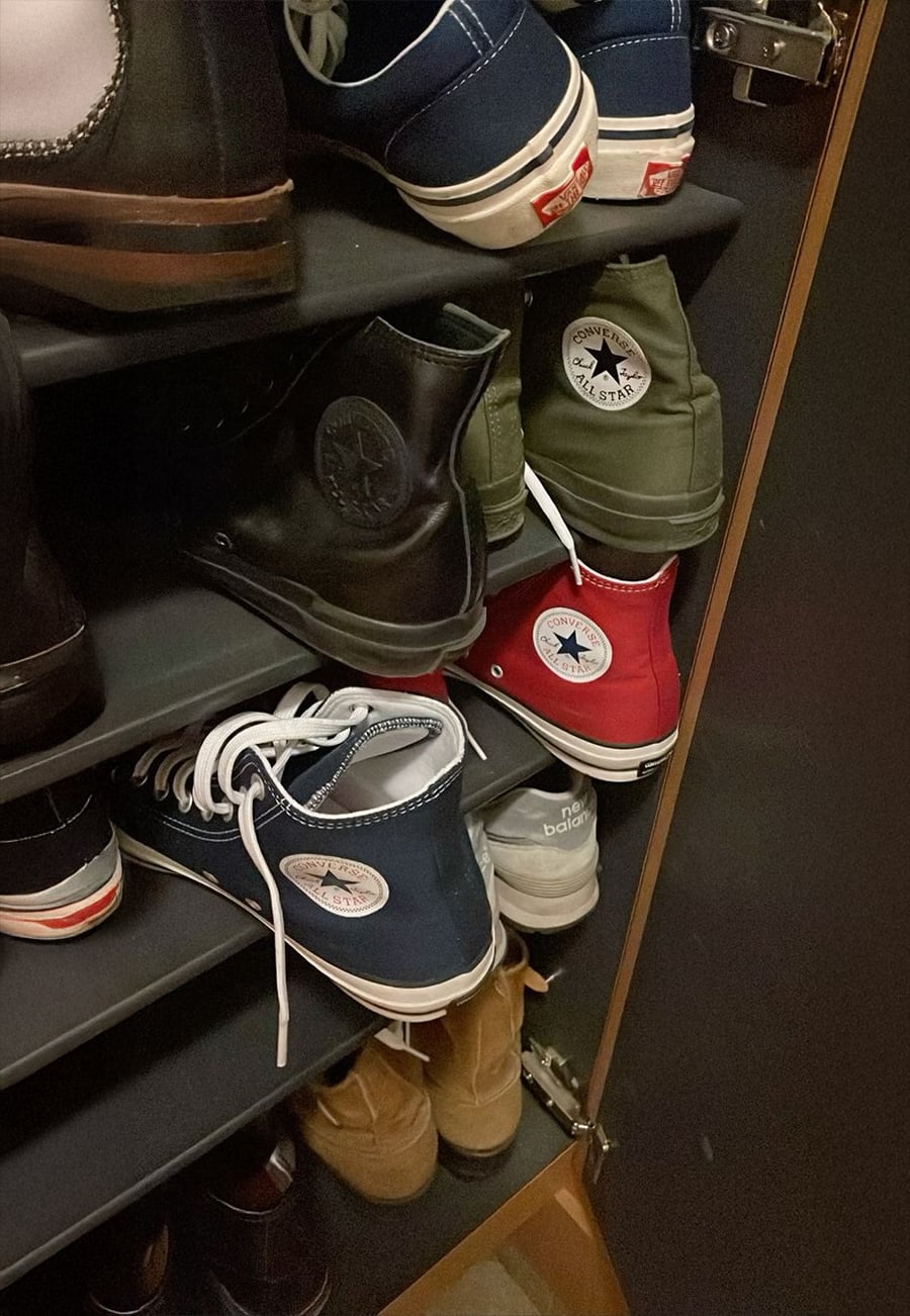Several pairs of Converse Chuck Taylor All Star shoes in black, green, blue, and red are neatly arranged on a black shoe rack inside a closet. Other types of shoes are visible on the shelves as well.