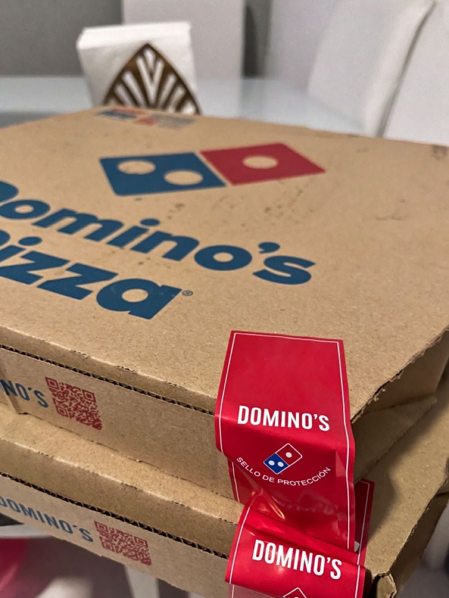 Two stacked Domino’s Pizza boxes with red security seals on a table. A chair with a decorative backrest and a folded napkin are visible in the background.