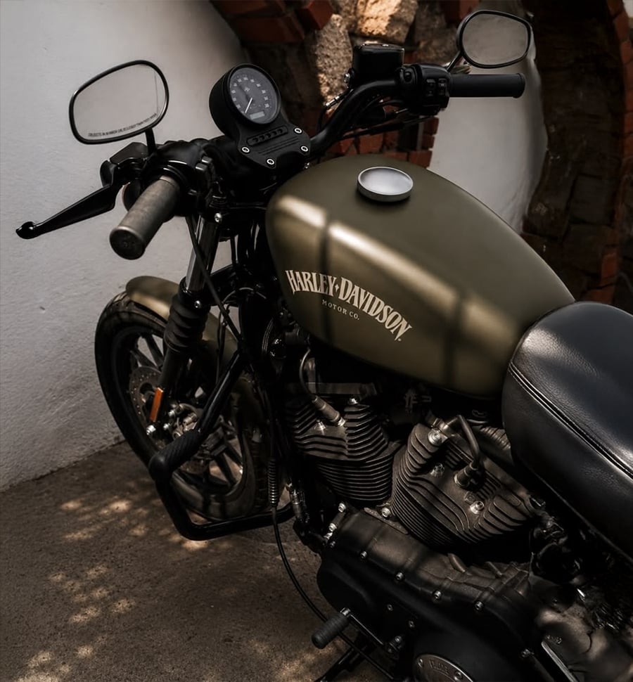 Matte green Harley-Davidson motorcycle parked indoors, sunlight casting shadows on the tank and engine, with brick and white walls in the background.