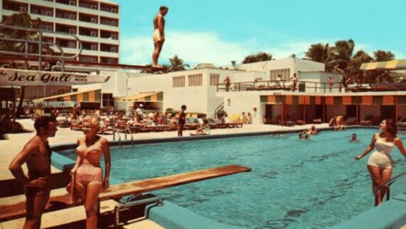 Vintage scene of people in swimsuits at a hotel pool; a man stands on a diving board, others relax poolside or swim, with a hotel building and palm trees in the background under a clear blue sky.