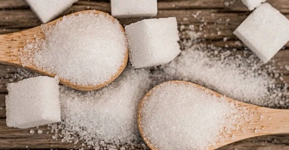 Two wooden spoons filled with granulated sugar are placed on a wooden surface surrounded by loose sugar crystals and several white sugar cubes.