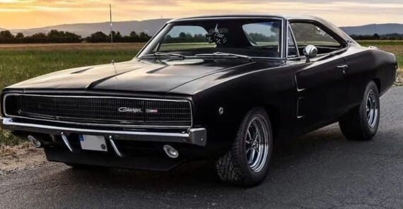 A black classic Dodge Charger muscle car is parked on a rural road at sunset, with fields and mountains in the background.