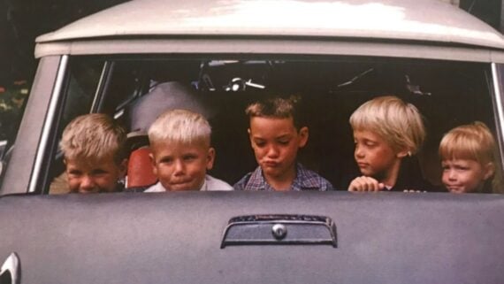 Five young children sit in the front seat of an old car, all making various facial expressions. Four have blond hair, and one has brown hair in pigtails. The car’s windshield and hood are visible.
