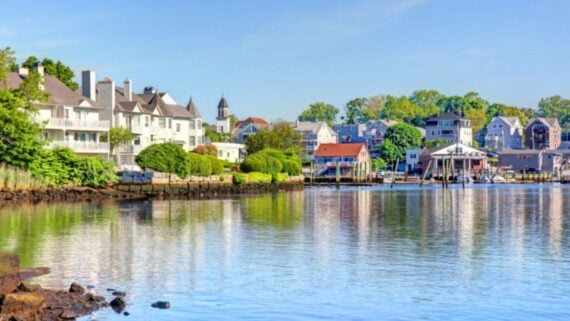 Waterfront houses with colorful facades and lush greenery line a calm river under a clear blue sky, reflecting in the water on a sunny day.