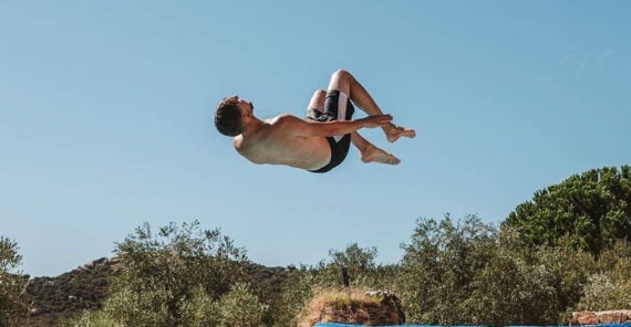 A person in swim trunks performs a mid-air flip against a clear blue sky, with trees and hills in the background.