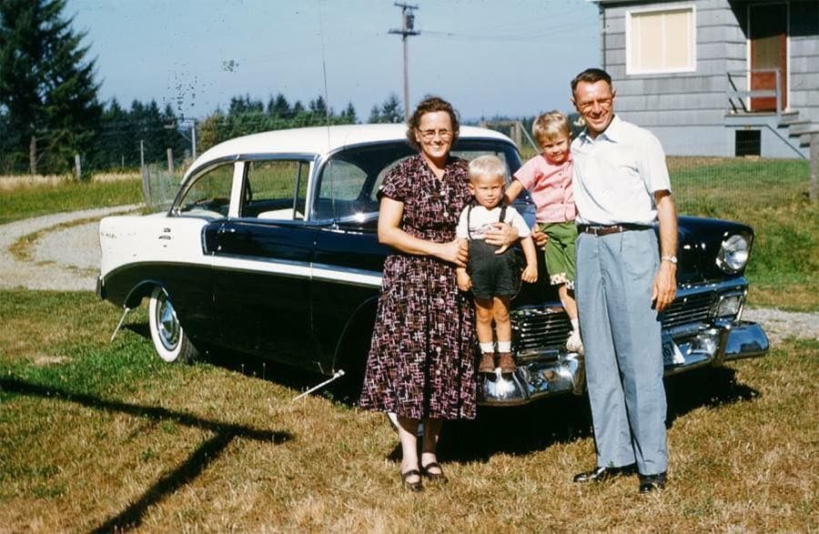 A family of four poses in front of a classic black and white car, smiling in a grassy yard with a house and trees in the background.
