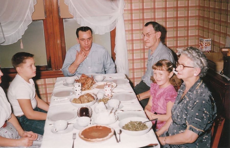 Six people, three adults and three children, sit around a dinner table set with food in a cozy, plaid-wallpapered dining room. The group appears to be enjoying a family meal together.
