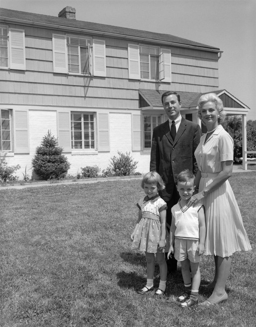 A smiling family of four, two adults and two children, stands on the lawn in front of a two-story house with shutters and siding, on a sunny day.