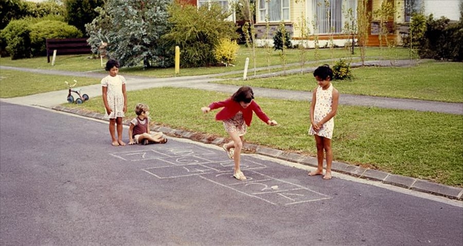 Four children play hopscotch on a suburban street, with one girl mid-jump while others watch. A scooter and green lawns are visible in the background near houses.