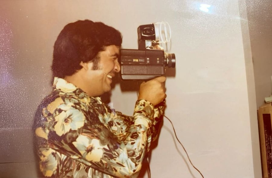 A man in a floral shirt smiles while holding a vintage video camera to his eye, standing indoors against a plain wall.