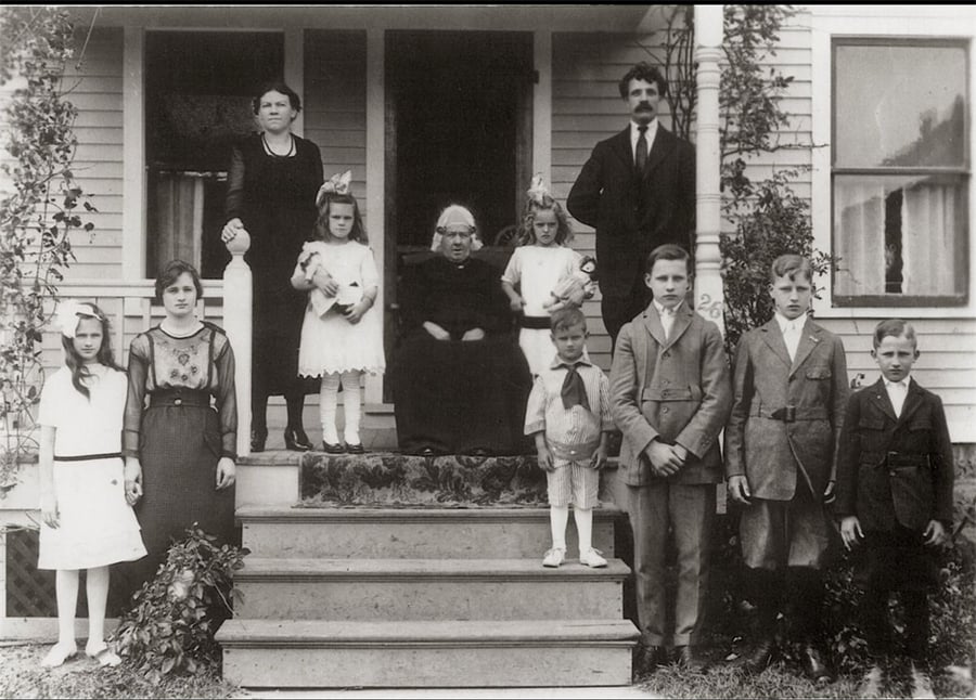 A black-and-white photo of eleven people, spanning three generations, pose on the steps of a house. There are five children, four women, and two men, all dressed in early 20th-century clothing.