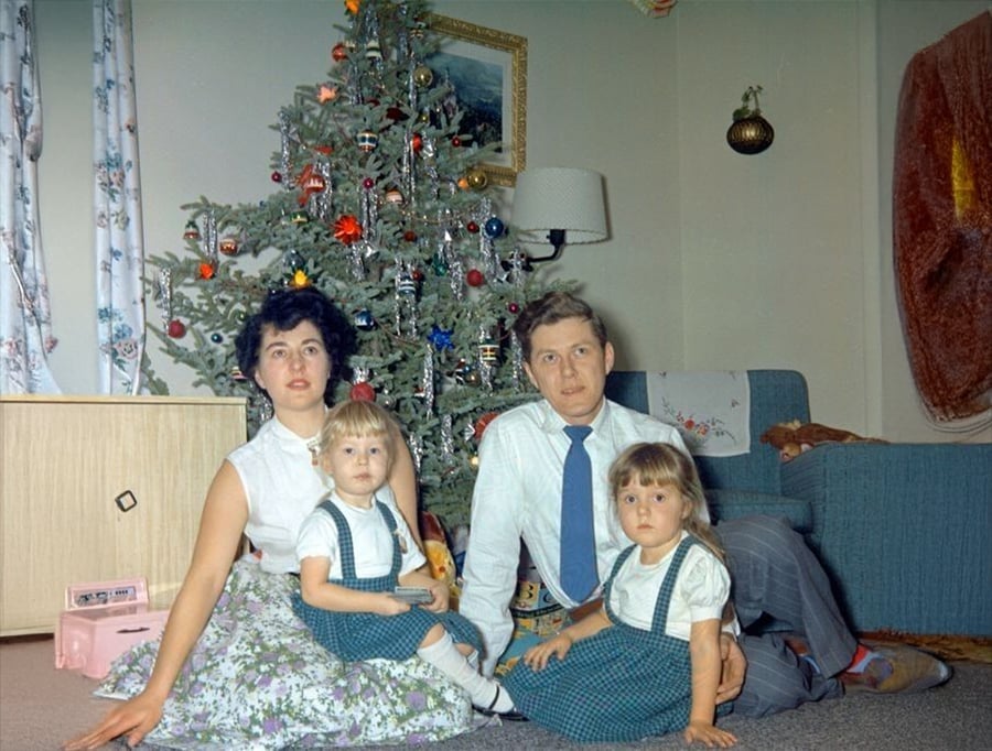 A family of four sits on the floor by a decorated Christmas tree, dressed in coordinated outfits. Two young girls are seated in front of their parents in a cozy living room with festive decorations.