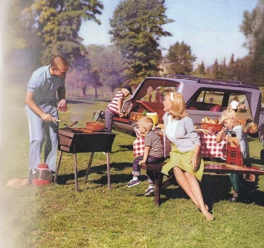 A family enjoys a picnic outdoors; a man grills food, two children sit at a bench with a woman, and a car with an open trunk is in the background. Trees and grass surround the area.