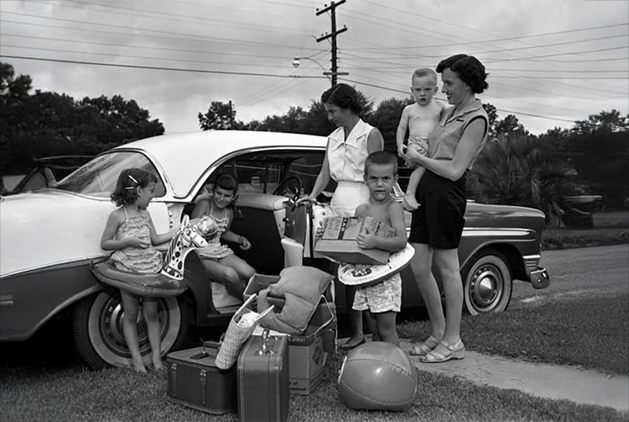 Two women and four children stand beside a vintage car packed with suitcases, beach toys, and floaties, preparing for a trip. The scene is outdoors on a grassy lawn under a cloudy sky.