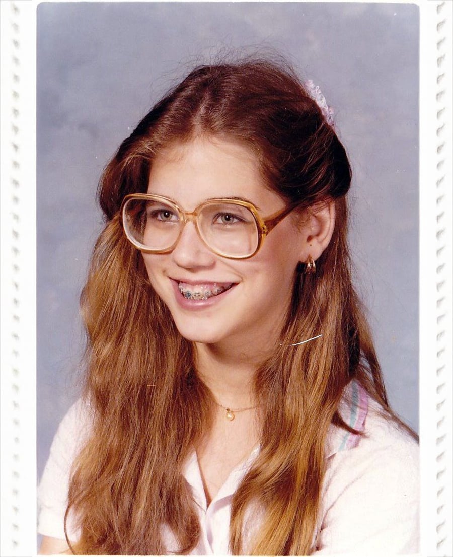 A teenage girl with long brown hair, large glasses, and braces smiles at the camera. She is wearing a light-colored shirt and a gold necklace, with a gray-blue background behind her.