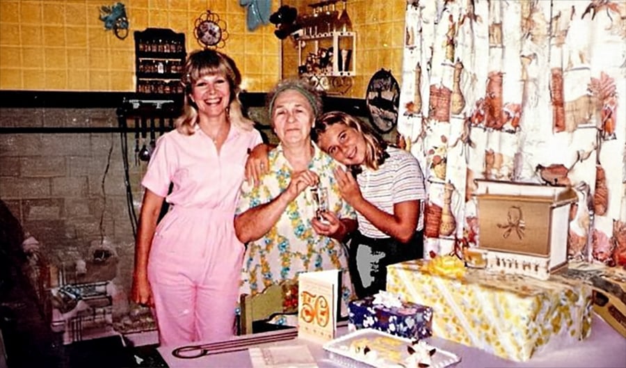 Three women stand together in a kitchen, smiling beside a decorated table with a “50” card, gifts, and cake, suggesting a celebration. The setting is colorful with patterned curtains and yellow tiles.