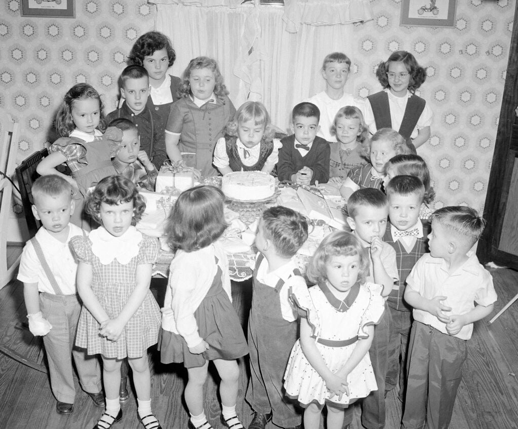 A group of young children in party clothes stand around a table with a birthday cake, posing for a photo in a decorated room with patterned wallpaper.