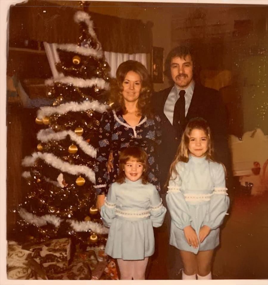 A family of four poses by a decorated Christmas tree. The parents stand behind their two young daughters, who are wearing matching blue dresses. Wrapped presents are visible under the tree.
