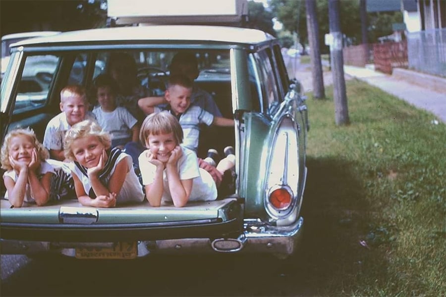 Seven smiling children lay in the open back of a green vintage station wagon parked on a grassy roadside. Some children rest their heads on their hands, enjoying a sunny day.